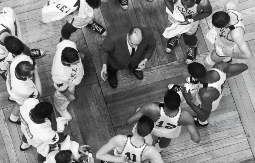 Black and white photo of a basketball team huddle. Players wearing jerseys, and a coach in a suit stand in a circle on a wooden court floor, discussing strategy. Jerseys display numbers and the word "Celtics.