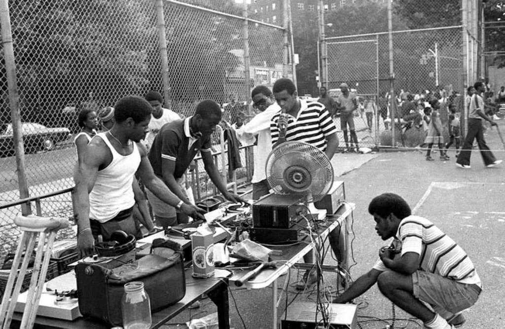 A group of people gathered around DJ equipment set up on a table in an outdoor urban park. Several individuals are adjusting the gear, while bystanders watch. A wire fence and various people are visible in the background.