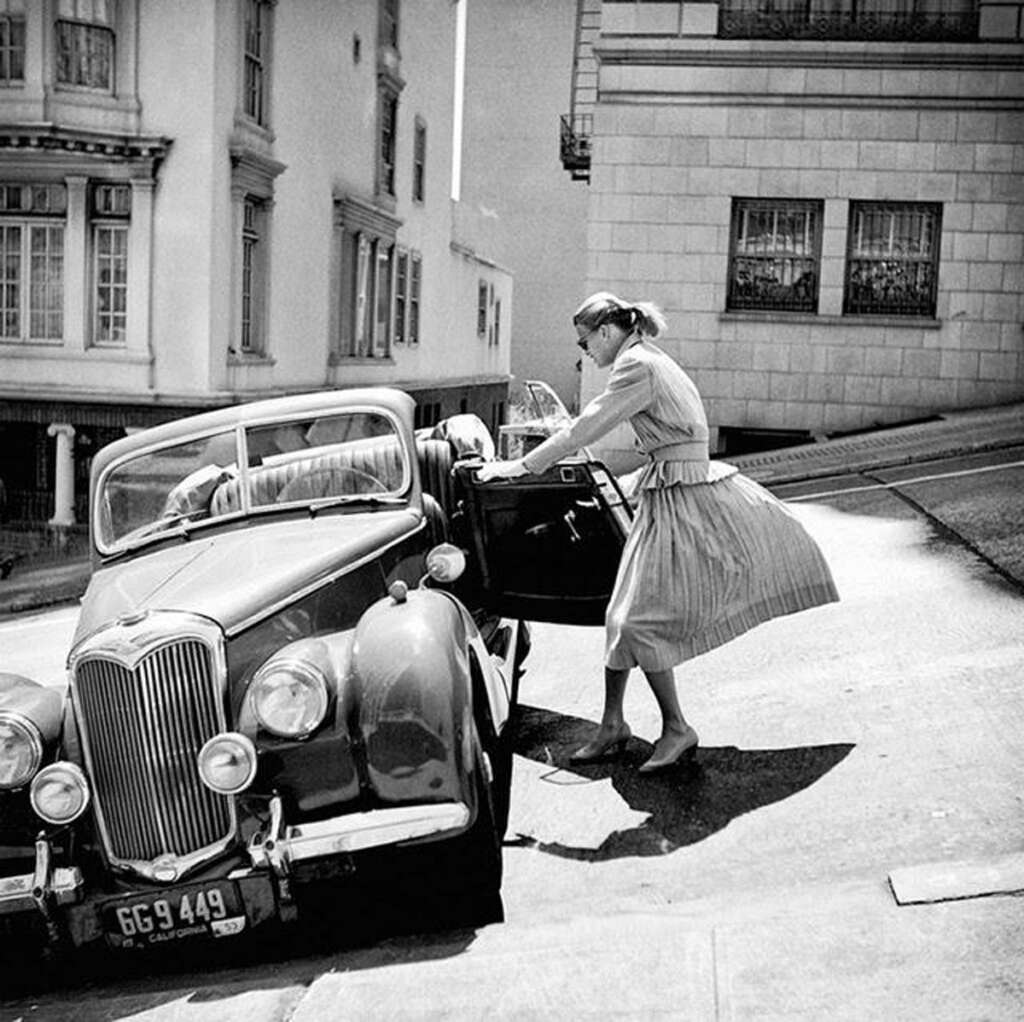 A woman in a dress struggles to open the door of a vintage convertible car parked on a steep street. Her skirt blows in the breeze, and buildings line the background. The scene is in black and white.