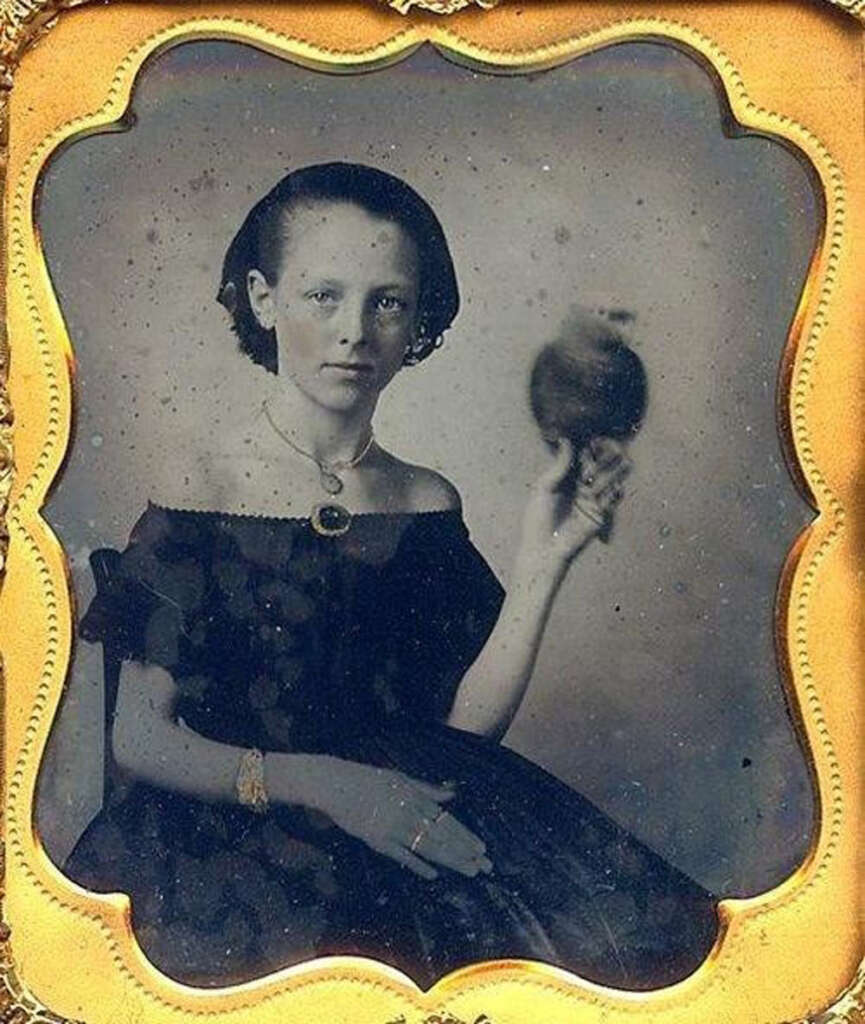 A young woman in a dark, off-the-shoulder dress holds a blurry spinning object in her right hand. She sits against a plain backdrop, gazing toward the camera. The image has an ornate gold frame around it.