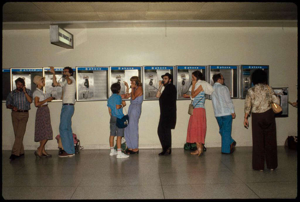 A line of people wait to use a row of payphones in a building. The group includes men and women in casual 1970s attire, with some talking on the phones, while others stand in line. A child with a backpack is also present.