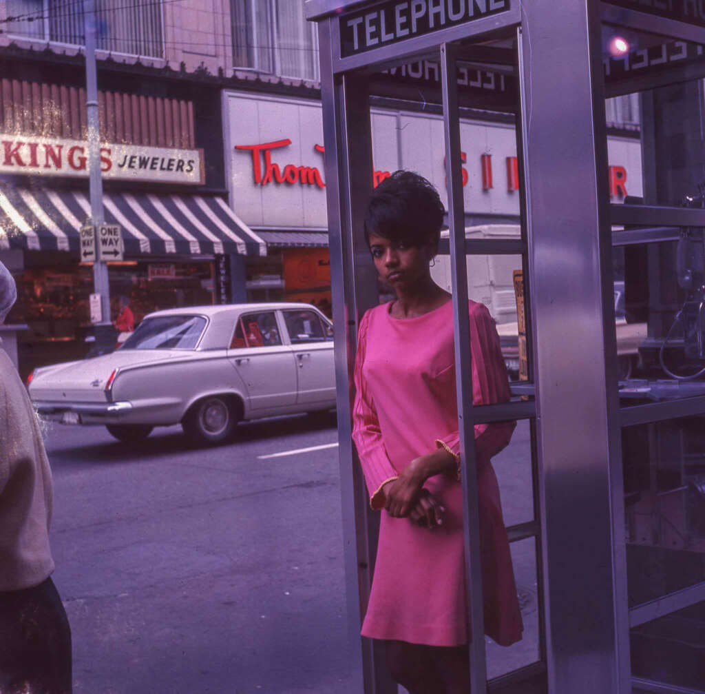 A woman in a pink dress stands inside a phone booth on a street. A vintage car and shops, including a jewelry store and bakery, are visible in the background.