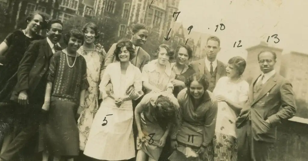 A vintage black and white photo of a group of men and women posing together. Some individuals are numbered on the image. They are dressed in 1920s-style clothing, standing in front of a building with large windows and brickwork.