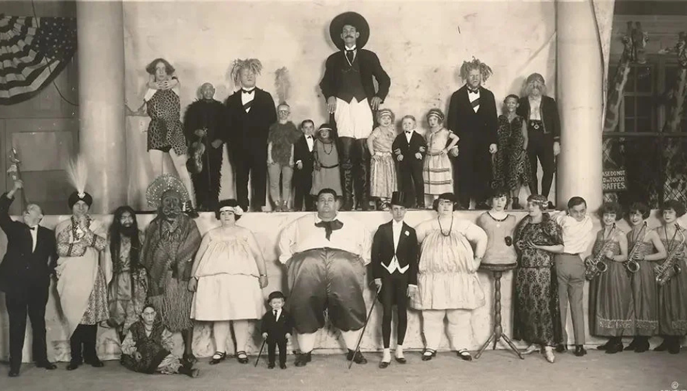 A vintage black and white photograph of a sideshow group featuring individuals with various physical characteristics and costumes. They are posed in two rows with some standing and others sitting, against a backdrop with an American flag on the left.