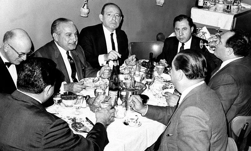 A group of six men in suits are sitting around a dining table engaged in conversation. The table is set with dishes, glasses, and wine bottles. The setting appears formal, possibly at a banquet or dinner event.