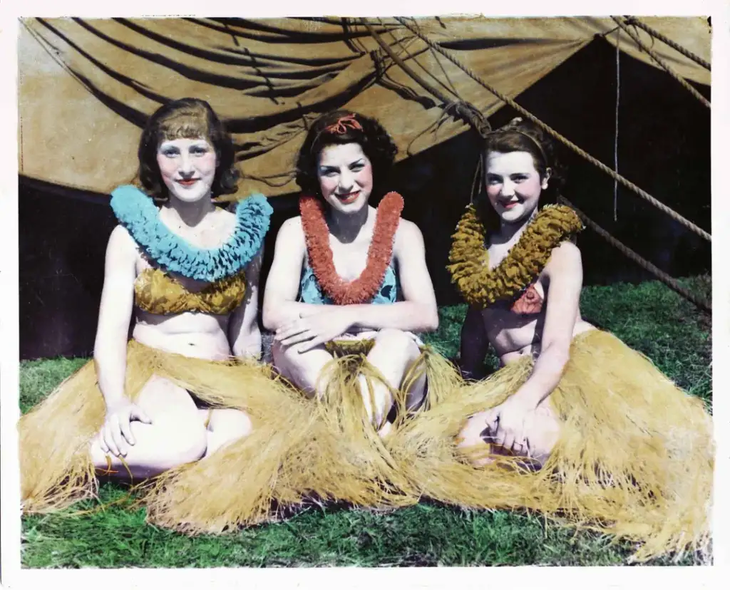 Three women sit on grass wearing hula skirts and colorful leis. They are dressed in tropical attire, with tents visible in the background. The scene suggests a festive or cultural event.