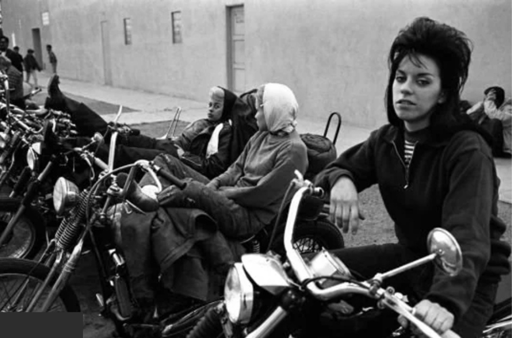 A black-and-white photo shows a group of people sitting on a row of motorcycles parked on the street. In the foreground, a woman with styled hair sits on one of the bikes, looking at the camera. Others are casually seated, facing right.