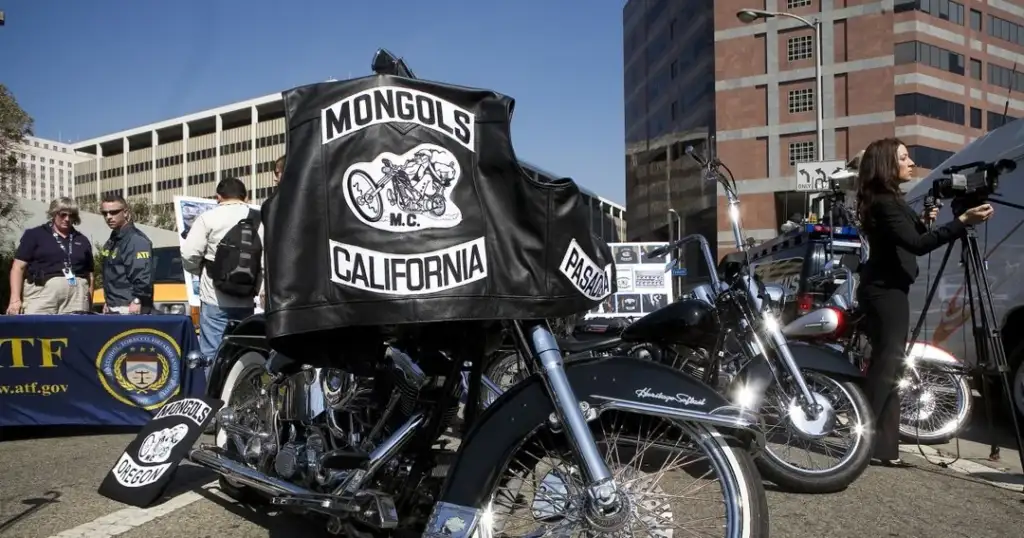 A motorcycle with "Mongols M.C. California" patches is displayed at an outdoor event. People are gathered around, some in conversation, and a woman is filming with a camera. Nearby are official banners and a few parked cars.