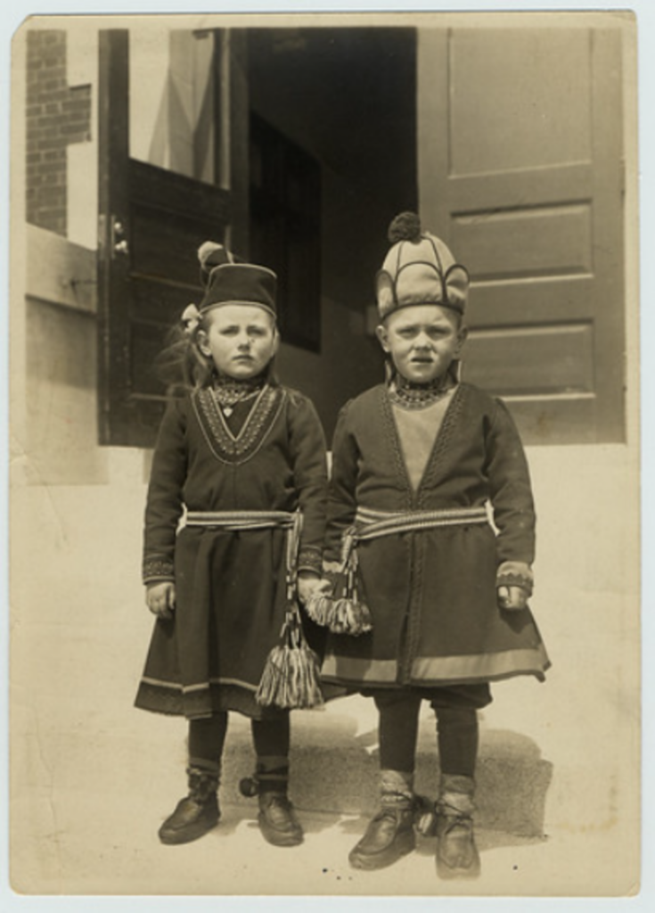 Two children stand holding hands, wearing traditional clothing with ornate embroidery and tassels. They both wear hats; the child's hat on the right is taller with a pom-pom. They are on steps in front of an open door. The photo is in sepia tone.