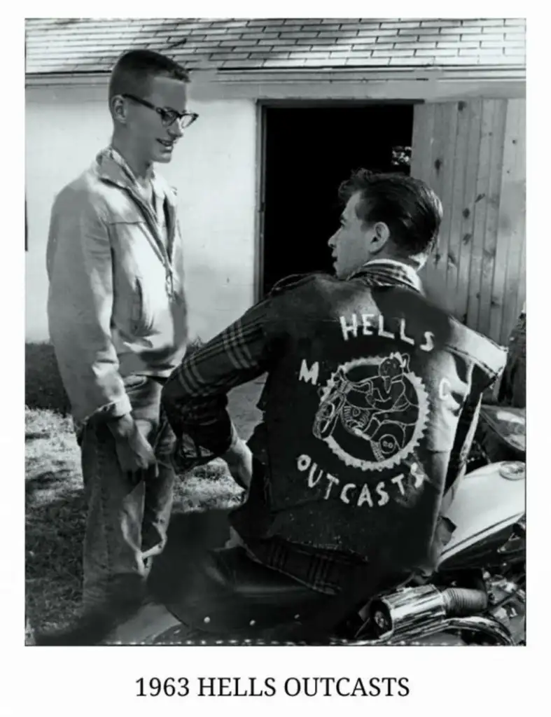 A man wearing a "Hells Outcasts MC" jacket sits on a motorcycle, facing another man with glasses and a jacket. They are outdoors near a building. The text "1963 HELLS OUTCASTS" is at the bottom of the image.