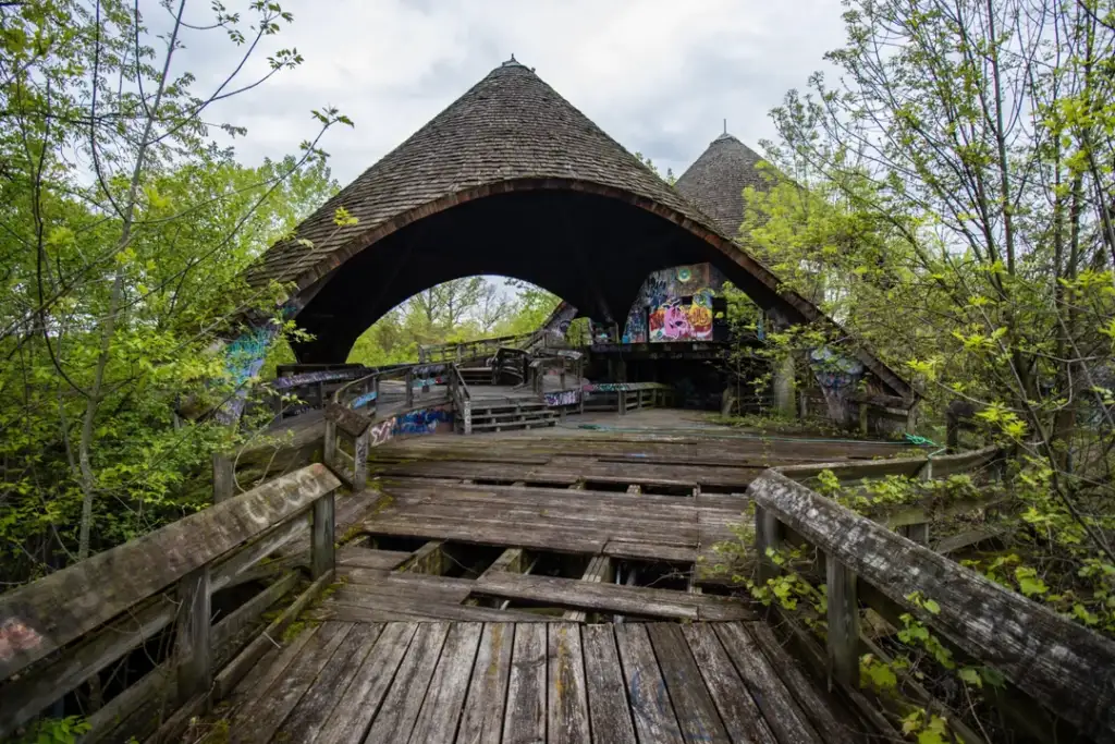 Abandoned, dilapidated wooden bridge leading to a large shingled structure, surrounded by dense greenery. The building features graffiti and has a steep, triangular roof, creating a moody atmosphere under a cloudy sky.