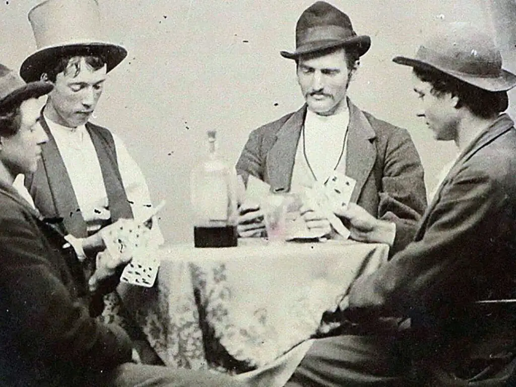 Vintage wild west photo of four men wearing hats are sitting around a small table playing cards. The table is covered with a patterned cloth, and there is a bottle in its center. The setting has a vintage feel, and the men appear focused on the game.