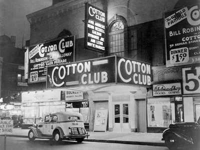 A black and white photo of the Cotton Club at night, illuminated by bright neon signs. Vintage cars are parked in front. Marquees advertise performances, including Bill Robinson and Cab Calloway. Surrounding buildings are visible in the background.