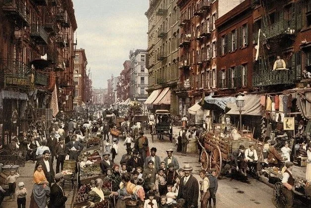 A bustling street market scene from the early 1900s shows a diverse crowd of people shopping and socializing. Horse-drawn carts, market stalls, and multi-story buildings line the street, capturing the vibrant atmosphere of the time. Part of a collection of old photos of New York City.