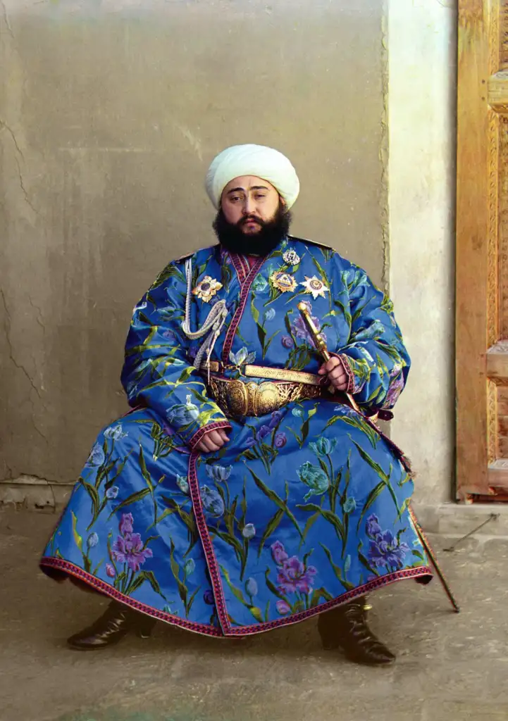 A man with a beard, wearing a luxurious blue robe adorned with floral patterns and a white turban, sits against a plain backdrop. He holds a sword and wears an ornate belt, exuding an air of nobility and authority.