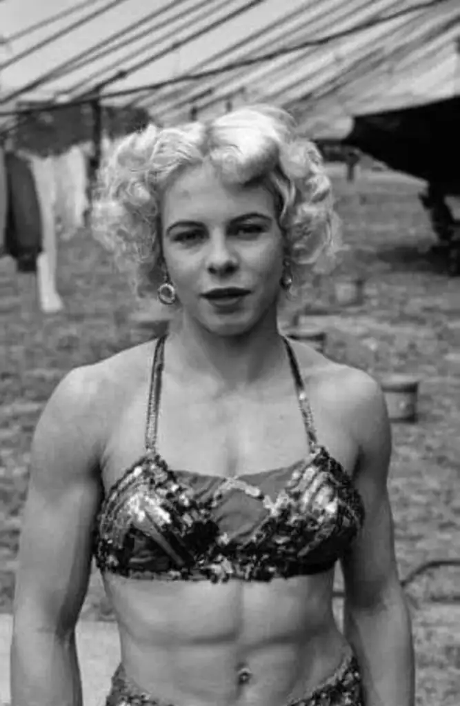 A woman with short curly hair stands outside, wearing a sequined top. The background shows a large striped tent and grassy area, suggesting a circus or fair setting.