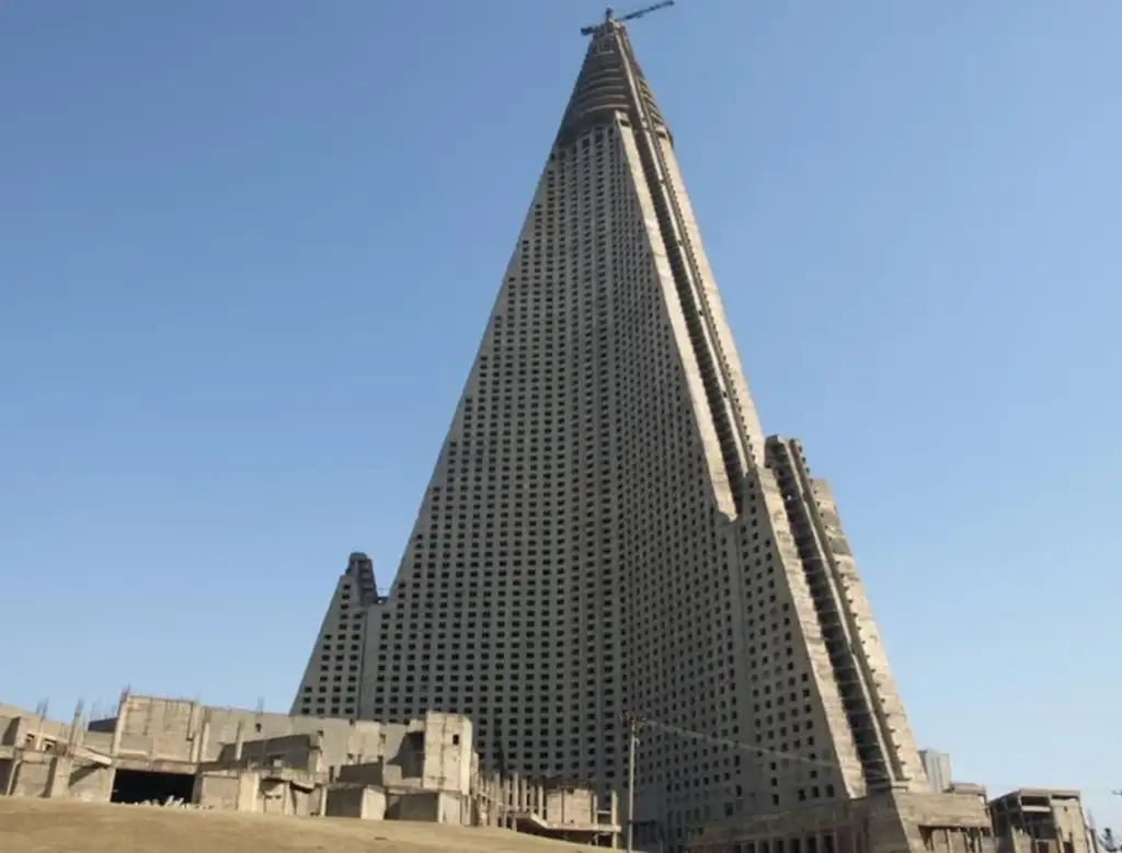 A tall, unfinished pyramid-shaped skyscraper under a clear blue sky, surrounded by construction debris at the base. A crane is positioned at the pointed top of the building. The structure appears to be made of concrete.