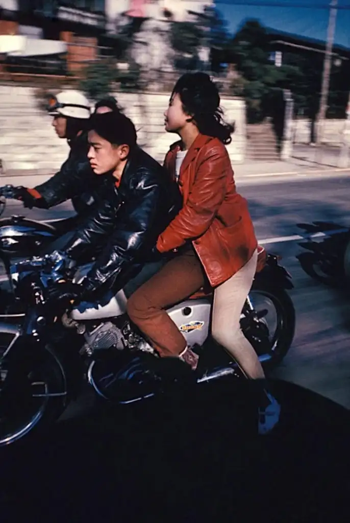 A young man and woman ride a motorcycle on a city street, both wearing leather jackets. The woman sits behind, holding onto the man. Other motorcyclists are blurred in motion, indicating speed. Buildings and a fence line the background.