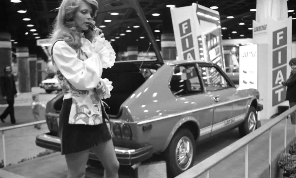 A woman in a patterned vest and blouse talks on a phone near a Fiat car with its hatch open, at an indoor auto show. There are signs and people in the background. The image is in black and white.