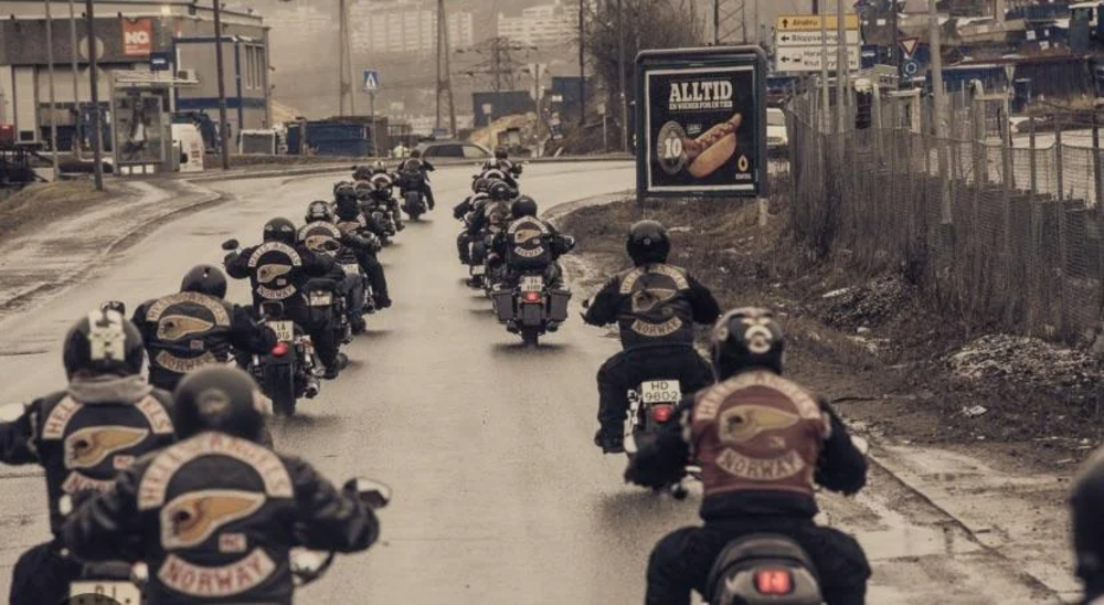 A group of motorcyclists wearing leather jackets with eagle patches ride down a wet road in an industrial area. Buildings and a billboard are visible in the background under a gray, overcast sky.