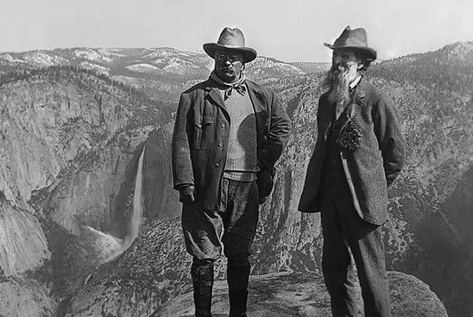 Two men wearing hats and outdoor clothing stand on a rocky ledge overlooking a valley. A waterfall cascades down a cliff in the distance, surrounded by forested mountains. The scene is in black and white, suggesting a historical context.