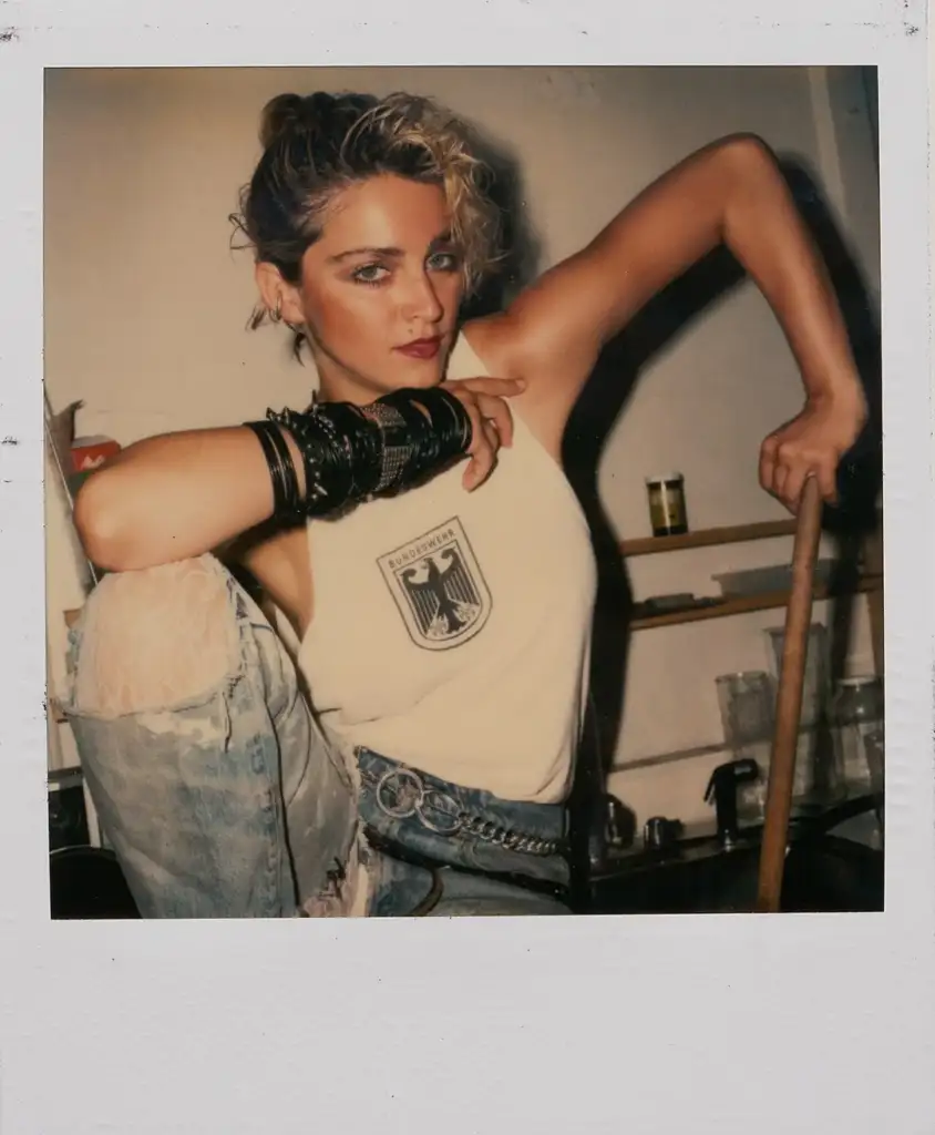 Woman with short blond hair and bold makeup wearing a sleeveless top and ripped jeans, poses indoors. She leans on one knee with an arm akimbo, wearing multiple bangles and a focused expression. Background includes shelves and kitchenware.