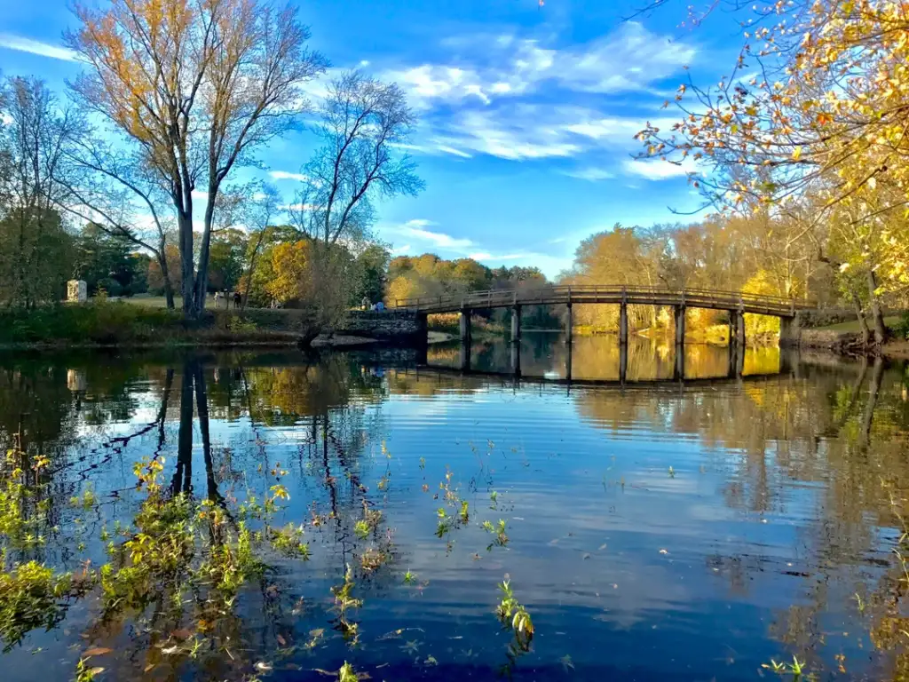 A serene scene of a wooden footbridge over a calm river, surrounded by trees in autumn colors. The clear blue sky with scattered clouds is reflected in the water, creating a picturesque landscape.