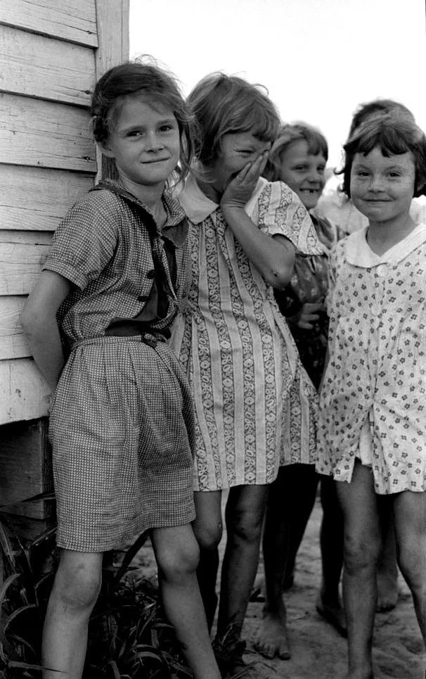 A black and white photo shows a group of five young girls standing by a wooden building. They wear patterned dresses with some barefoot. One girl leans casually against the wall, another giggles, and others smile at the camera.