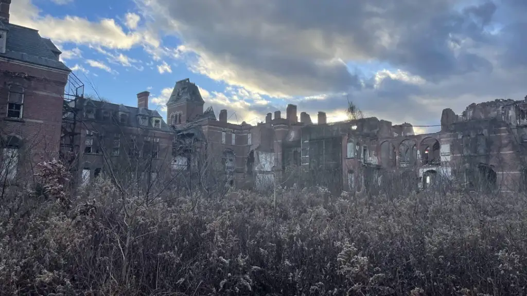 Abandoned, dilapidated brick building with collapsed roof and missing windows, surrounded by overgrown vegetation. The sky is cloudy with patches of blue visible. The scene conveys a sense of decay and neglect.