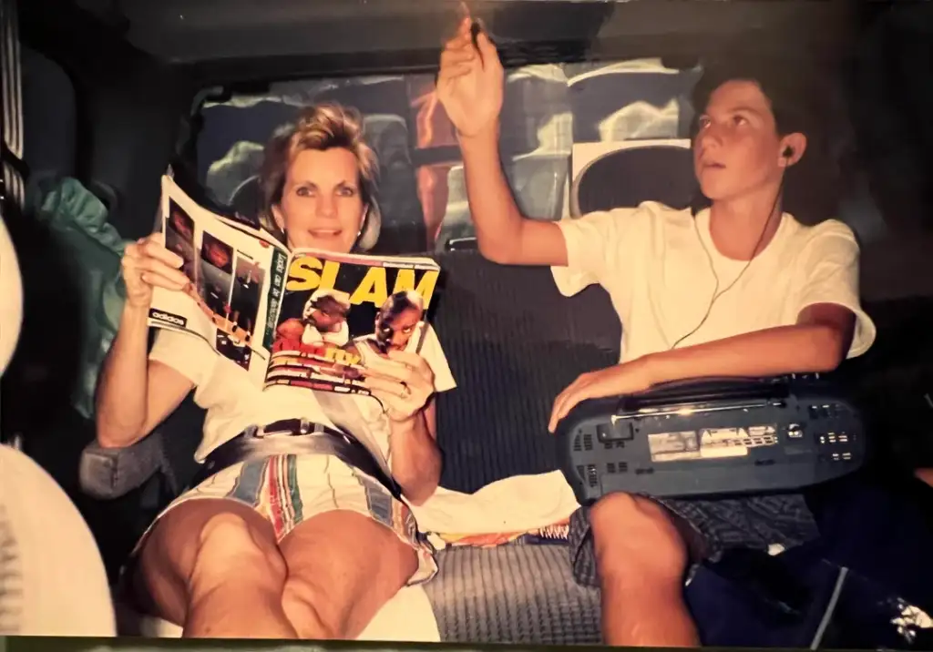 A woman and a young boy are sitting in a car. The woman is reading a magazine titled "SLAM" and wearing headphones, while the boy, also with headphones, is holding a boombox and reaching up with one hand. Both appear relaxed and casual.