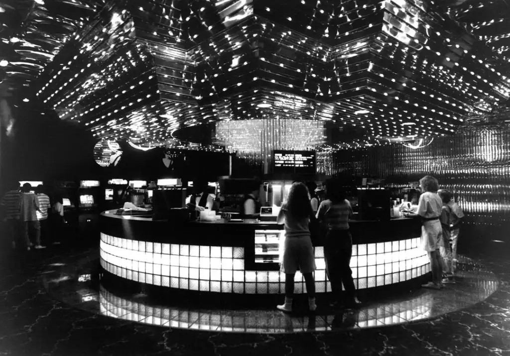 Black and white image of a retro arcade with a central, rounded counter. Ceiling covered in reflective tiles and bright lights. People are gathered around the counter, and arcade machines are visible in the background.