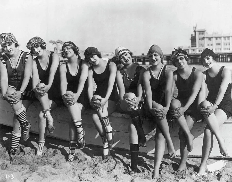 A vintage black-and-white photo shows eight women wearing swimsuits and swim caps sitting on a wooden structure at the beach. They are smiling and leaning playfully, with a pier and a large building visible in the background.