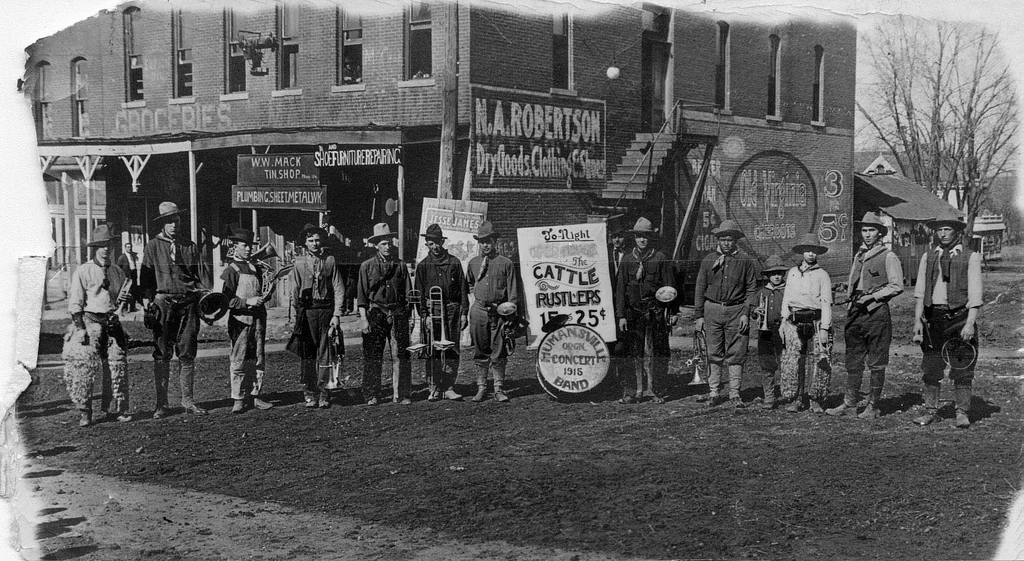 A vintage photo shows a band of 14 men holding musical instruments, including trombones and saxophones, standing on a dirt road. A sign advertises a performance. Behind them is a brick building with shop signs and a staircase.