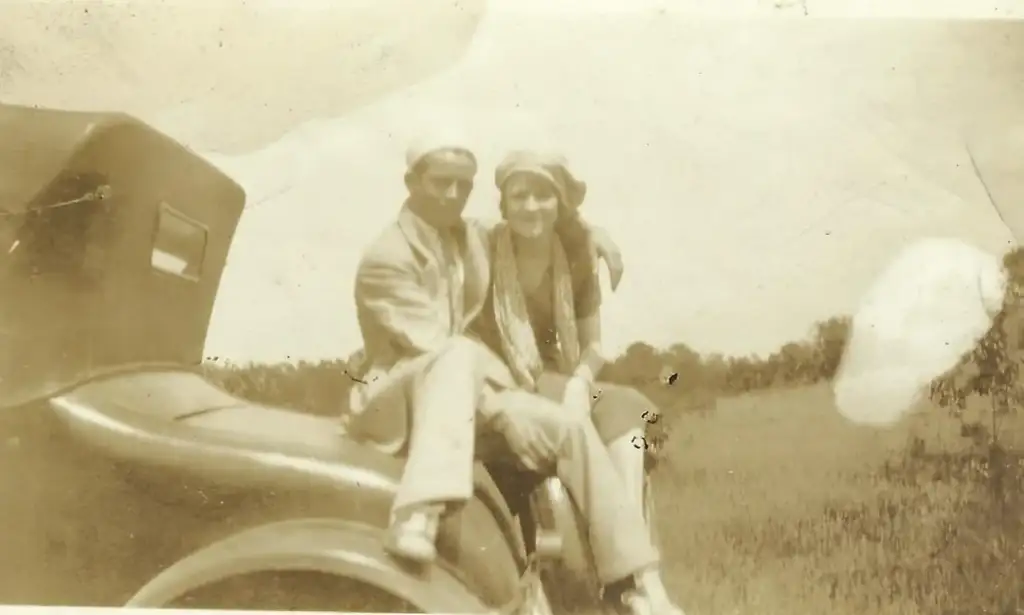 A sepia-toned vintage photo of a couple sitting on the front of an old car. They are wearing early 20th-century clothing, with the woman wearing a headscarf. A grassy field and trees are visible in the background.