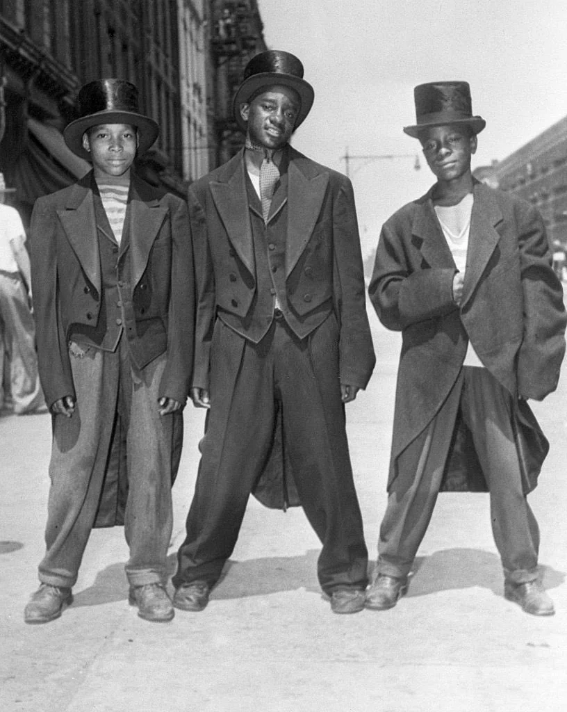 Three boys stand confidently on a street, each wearing oversized formal coats and top hats. The black-and-white photo suggests a historical setting, possibly from the early to mid-20th century, with buildings visible in the background.