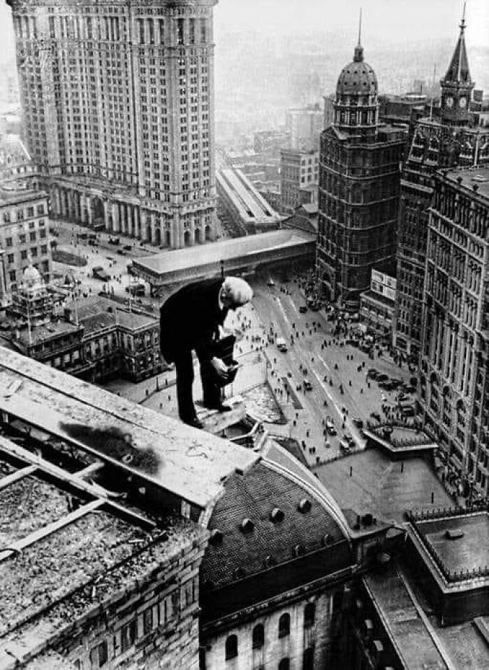 A person crouches on the edge of a tall building overlooking a bustling cityscape below, with historical architecture, including a clock tower and large crowds on the streets. The scene appears to be in black and white.