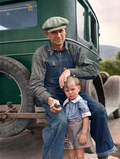 A man in a cap, denim shirt, and overalls sits on the bumper of a green vehicle, holding a pipe. He gently rests his hand on the head of a young boy in a blue shirt and shorts standing beside him. The background shows greenery.