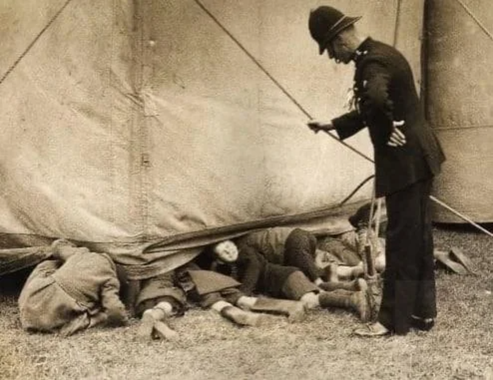 A vintage photograph showing a police officer looking at several children who are peeking under a large tent. The children are lying on the ground, partially under the tent fabric, while the officer stands watching them with hands on hips.