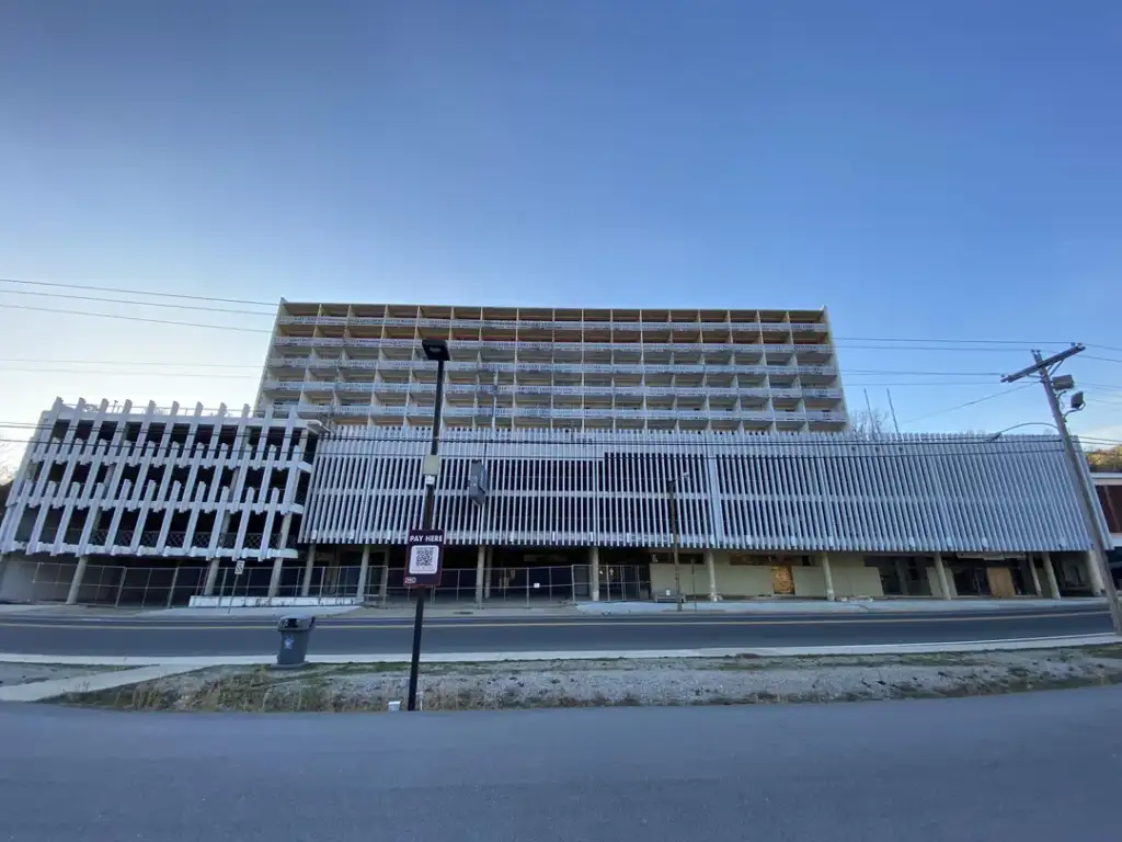 A partially constructed building with exposed concrete floors is seen under a clear blue sky. The facade features vertical beams. Construction materials and a sign are visible on the ground in front of the structure.