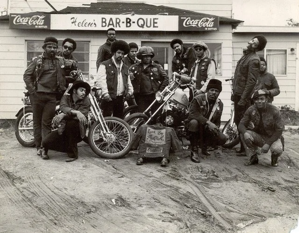 A group of bikers posing in front of Helen's Bar-B-Que. They are dressed in leather jackets and standing with their motorcycles. A Coca-Cola sign is visible on the building's facade. The ground is dirt, and the atmosphere is casual and lively.