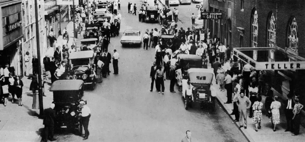 A bustling 1930s street scene with numerous people walking and standing along the sidewalks. Vintage cars are parked on both sides of the street. Signs for "West Virginia Grill" and "Teller" are visible among the brick buildings.