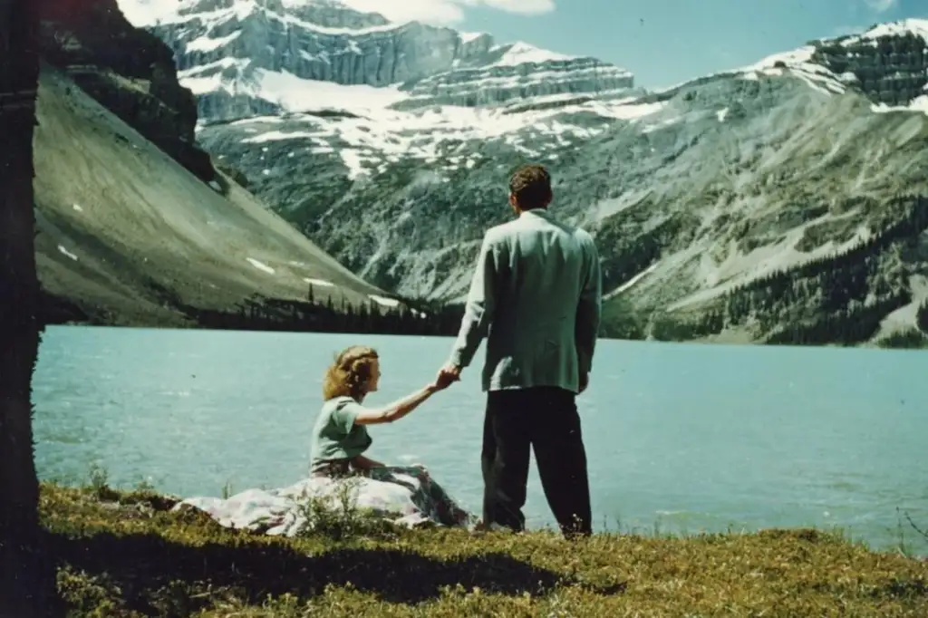 A couple is sitting near a serene mountain lake. The woman is seated on the grass, reaching out, while the man stands holding her hand. Snow-capped mountains tower in the background under a clear blue sky.