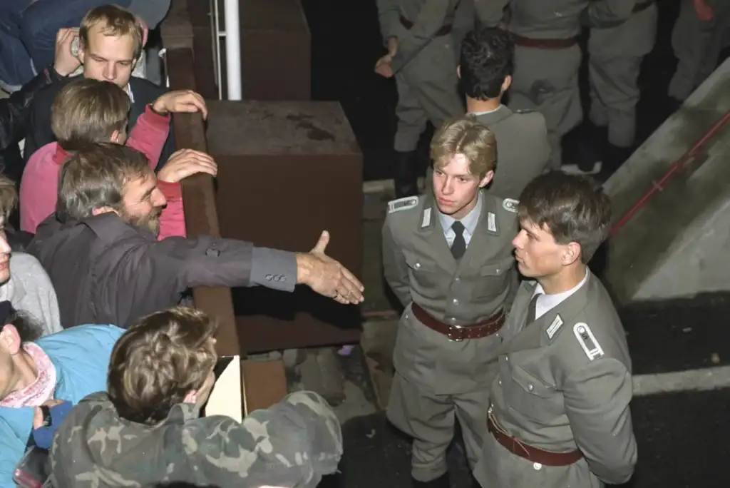 A man extends his hand over a wall towards two uniformed guards, who stand with their arms crossed. Several people gather on the other side, watching the interaction. The scene is dimly lit, suggesting it might be evening or nighttime.