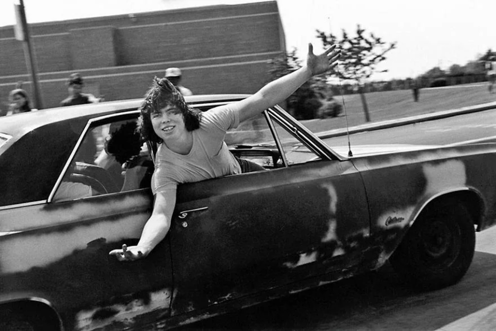 A person with shaggy hair leans out of the passenger side of a moving car, smiling with one arm extended. Another person drives. The car appears weathered, and the scene is outdoors near a brick building and a few trees.