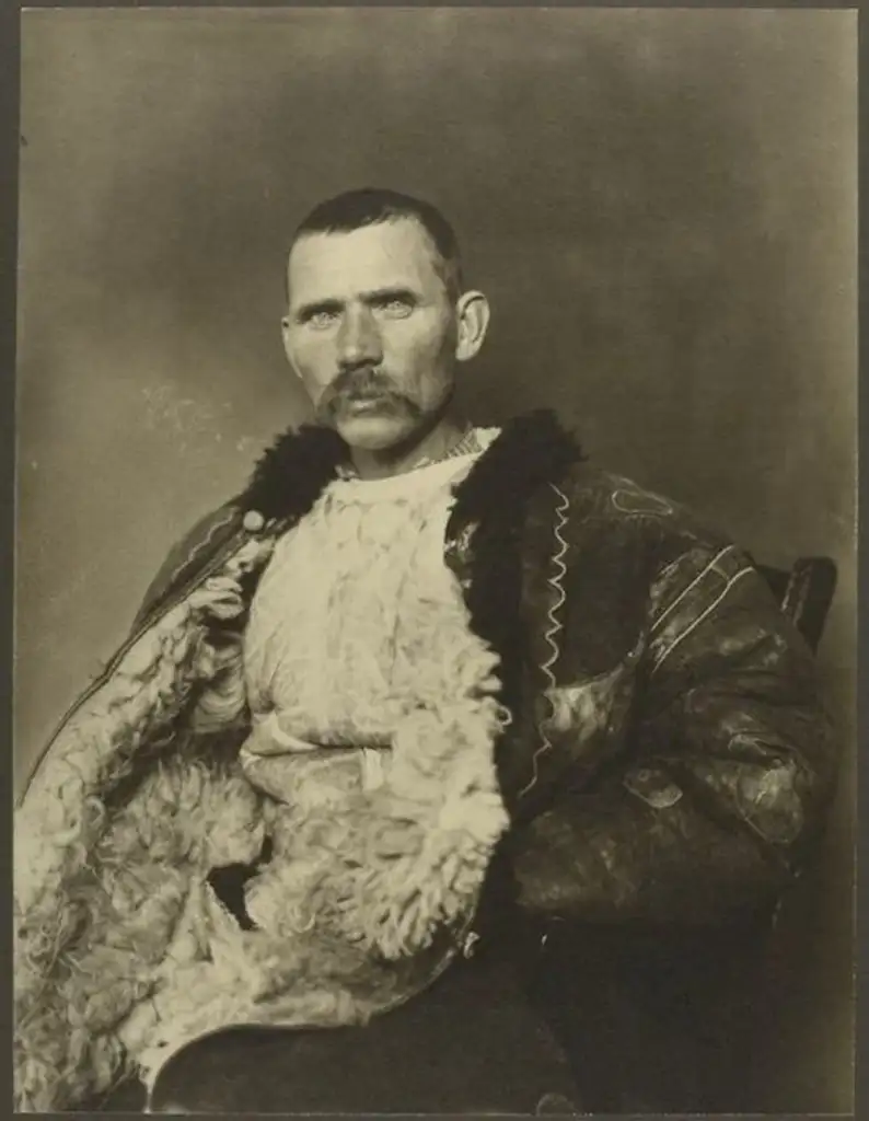 A man with a mustache, wearing a thick, fur-lined jacket, sits on a chair. He has a serious expression and is looking directly at the camera. The image is sepia-toned, giving it a vintage appearance.