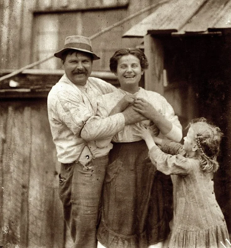 A sepia-toned vintage photo shows a smiling couple holding hands, with a young girl reaching up to them. The man wears a hat and the woman is dressed in a long skirt. They stand in front of a rustic wooden building.