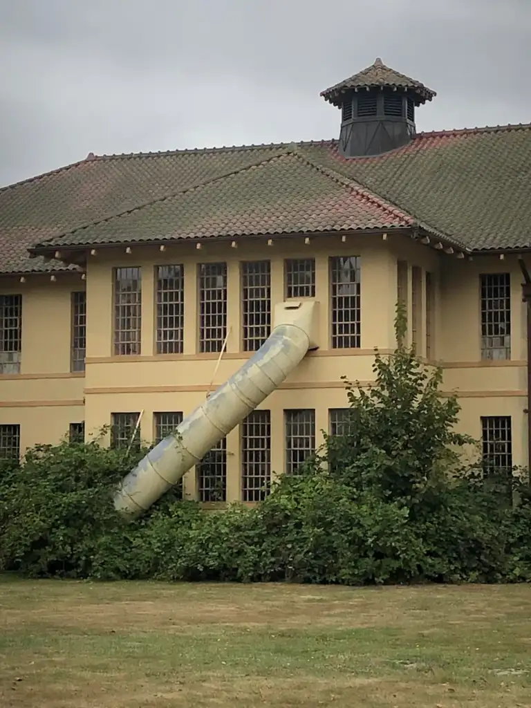 A beige stucco building with a brown tiled roof features a large cylindrical slide emerging from a second-story window, curving down into surrounding bushes. The building has numerous tall, rectangular windows and an octagonal roof structure.