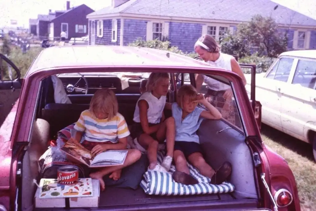 A group of four children sit in the back of a red station wagon. Two kids read a magazine, another looks outside, and the fourth gazes ahead. A woman stands nearby outside. The scene is set in a residential area with houses in the background.