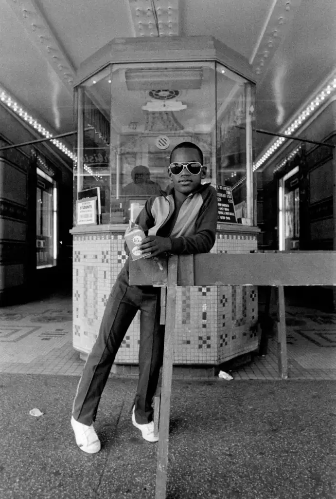 A young boy wearing sunglasses and a tracksuit leans casually against a barrier in front of a vintage theater ticket booth. He holds a drink cup and strikes a confident pose under the marquee lights.