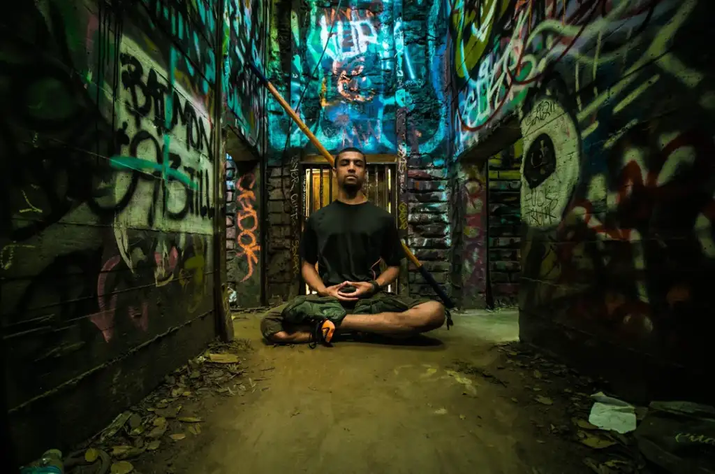 A person sits cross-legged meditating in a dimly lit, graffiti-covered passageway. Brightly colored street art decorates the walls, creating a vibrant and eclectic backdrop. The ground is strewn with leaves and debris, adding to the gritty urban atmosphere.